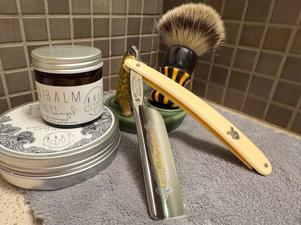 On a gray microfiber towel a straight razor with blade open about 45 degrees leans next to a bottle of aftershave and a jar of shave soap. A shave brush rests bristles-up in a green Wade ceramic pipe rest that features a brown Cairn Terrier.