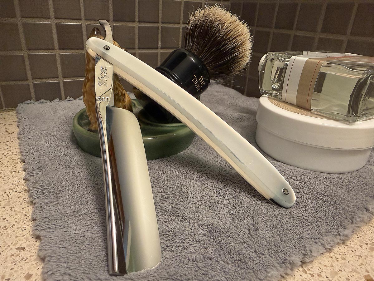 On a gray microfiber towel a straight razor with blade open about 45 degrees leans next to a bottle of aftershave and a jar of shave soap. A shave brush rests bristles-up in a green Wade ceramic pipe rest that features a brown Cairn Terrier.