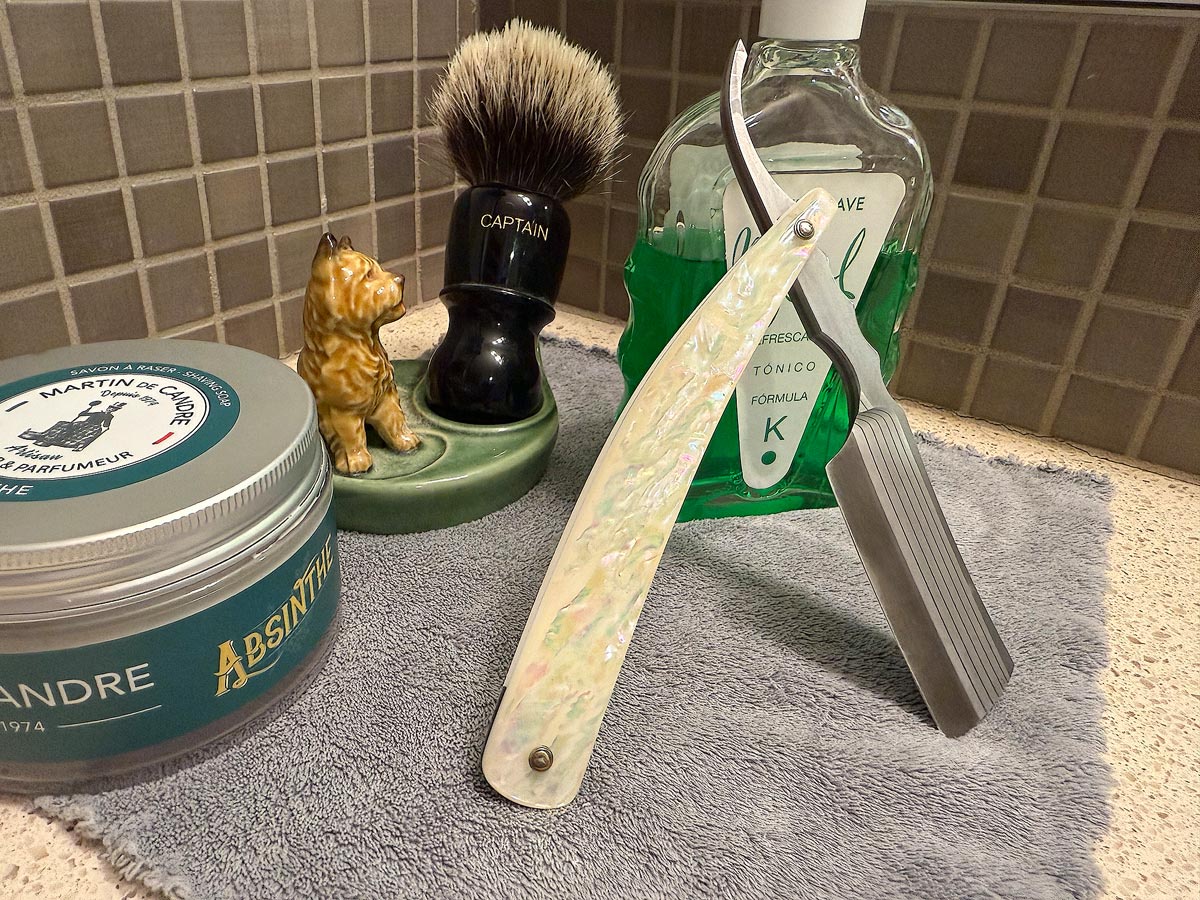 On a gray microfiber towel a straight razor with blade open about 45 degrees leans next to a bottle of aftershave and a jar of shave soap. A shave brush rests bristles-up in a green Wade ceramic pipe rest that features a brown Cairn Terrier.