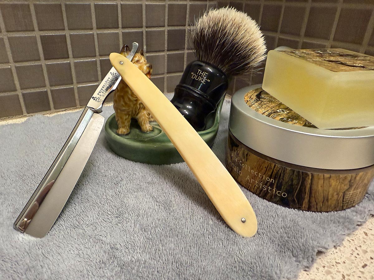 On a gray microfiber towel a straight razor with blade open about 45 degrees leans next to a bottle of aftershave and a jar of shave soap. A shave brush rests bristles-up in a green Wade ceramic pipe rest that features a brown Cairn Terrier.