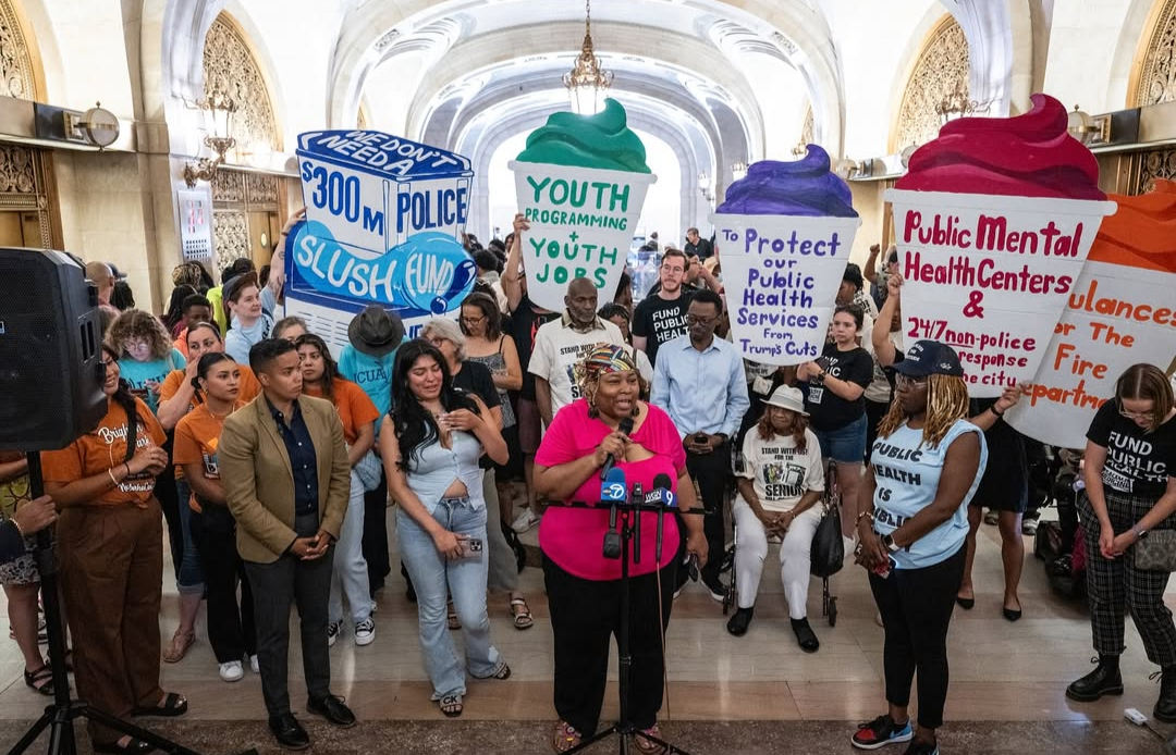 A group of protestors hold a press conference in an ornate hall, holding large paper cutouts of slushie drinks with writing on them.