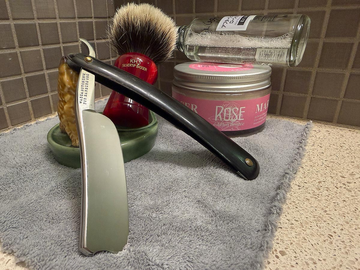 On a gray microfiber towel a straight razor with blade open about 45 degrees leans next to a bottle of aftershave and a jar of shave soap. A shave brush rests bristles-up in a green Wade ceramic pipe rest that features a brown Cairn Terrier.
