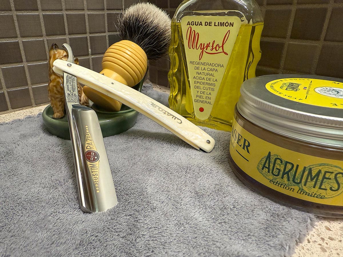 On a gray microfiber towel a straight razor with blade open about 45 degrees leans next to a bottle of aftershave and a jar of shave soap. A shave brush rests bristles-up in a green Wade ceramic pipe rest that features a brown Cairn Terrier.