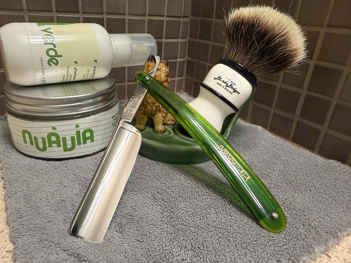 On a gray microfiber towel a straight razor with blade open about 45 degrees leans next to a bottle of aftershave and a jar of shave soap. A shave brush rests bristles-up in a green Wade ceramic pipe rest that features a brown Cairn Terrier.