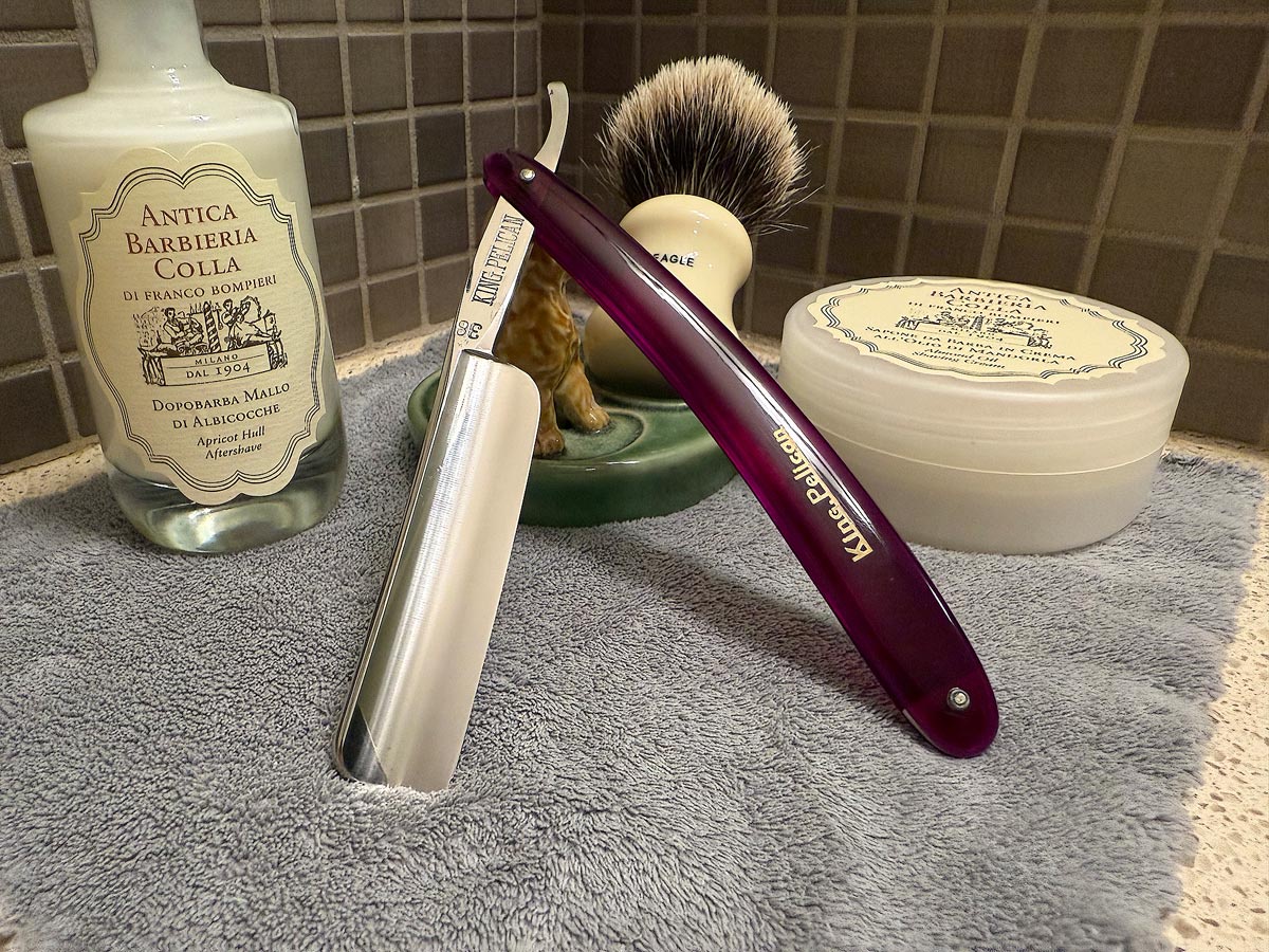 On a gray microfiber towel a straight razor with blade open about 45 degrees leans next to a bottle of aftershave and a jar of shave soap. A shave brush rests bristles-up in a green Wade ceramic pipe rest that features a brown Cairn Terrier.