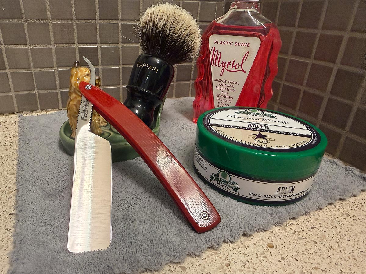 On a gray microfiber towel a straight razor with blade open about 45 degrees leans next to a bottle of aftershave and a jar of shave soap. A shave brush rests bristles-up in a green Wade ceramic pipe rest that features a brown Cairn Terrier.