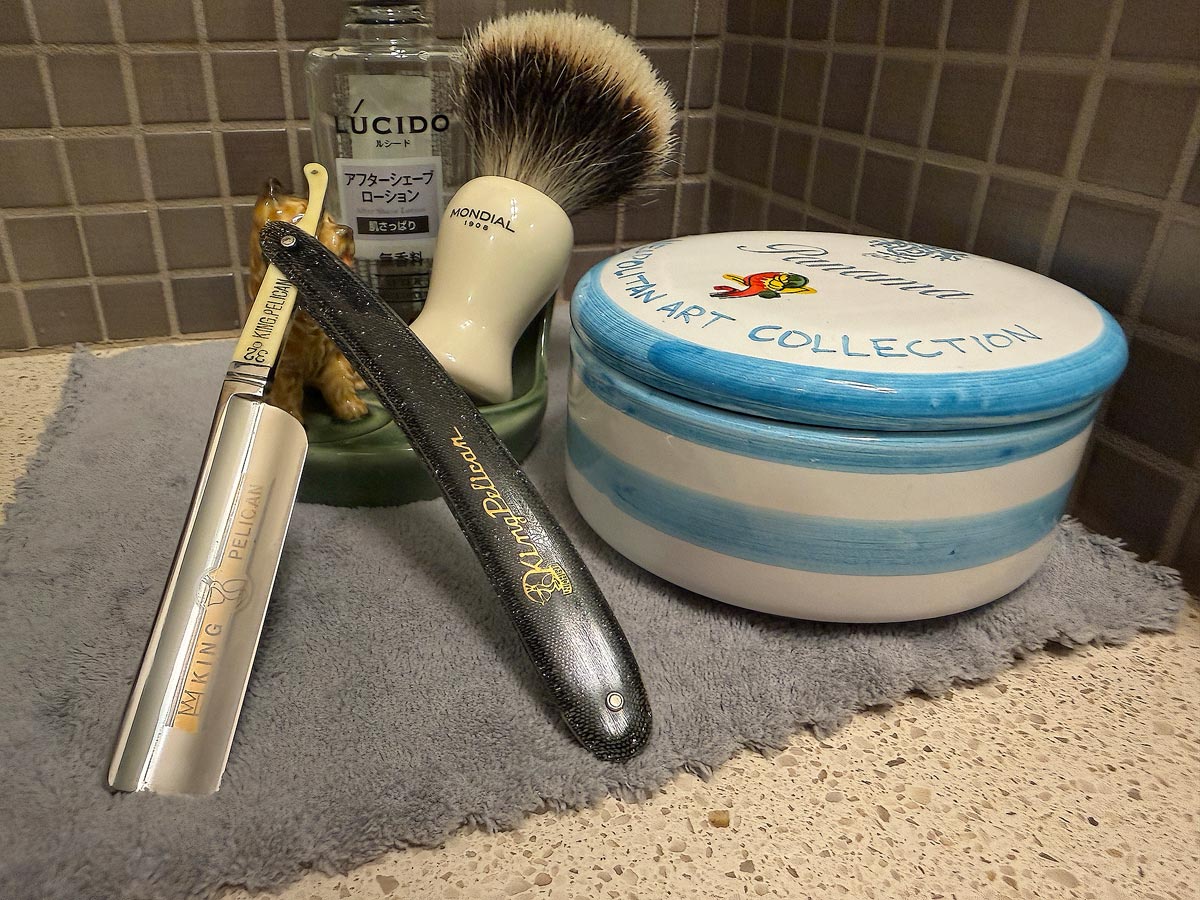 On a gray microfiber towel a straight razor with blade open about 45 degrees leans next to a bottle of aftershave and a jar of shave soap. A shave brush rests bristles-up in a green Wade ceramic pipe rest that features a brown Cairn Terrier.