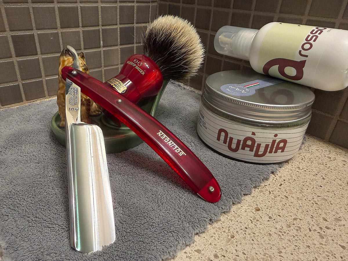 On a gray microfiber towel a straight razor with blade open about 45 degrees leans next to a bottle of aftershave and a jar of shave soap. A shave brush rests bristles-up in a green Wade ceramic pipe rest that features a brown Cairn Terrier.