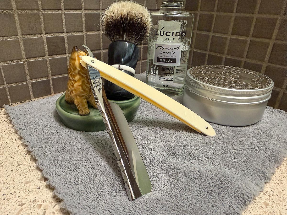 On a gray microfiber towel a straight razor with blade open about 45 degrees leans next to a bottle of aftershave and a jar of shave soap. A shave brush rests bristles-up in a green Wade ceramic pipe rest that features a brown Cairn Terrier.