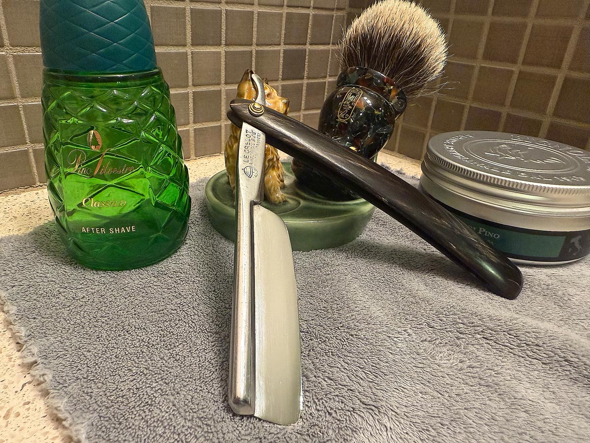 On a gray microfiber towel a straight razor with blade open about 45 degrees leans next to a bottle of aftershave and a jar of shave soap. A shave brush rests bristles-up in a green Wade ceramic pipe rest that features a brown Cairn Terrier.