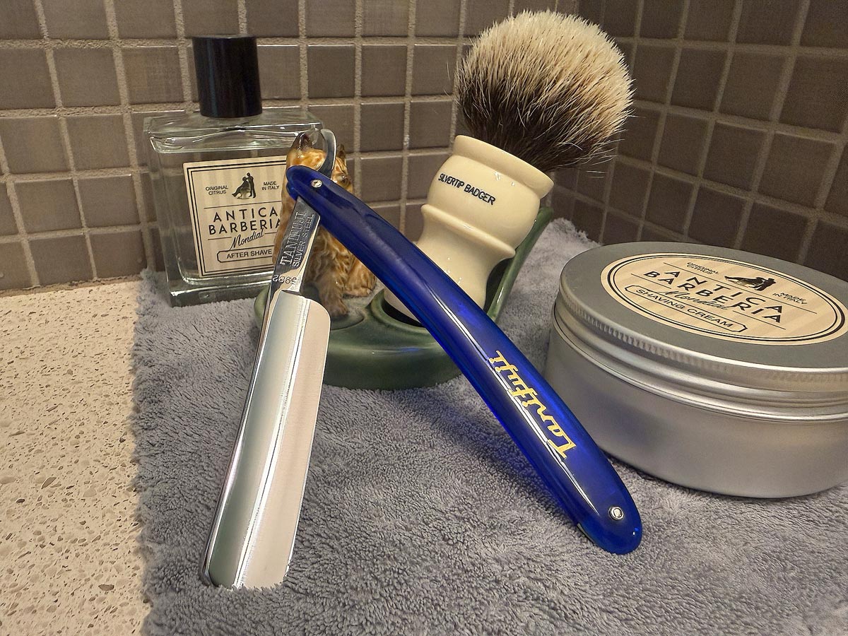 On a gray microfiber towel a straight razor with blade open about 45 degrees leans next to a bottle of aftershave and a jar of shave soap. A shave brush rests bristles-up in a green Wade ceramic pipe rest that features a brown Cairn Terrier.