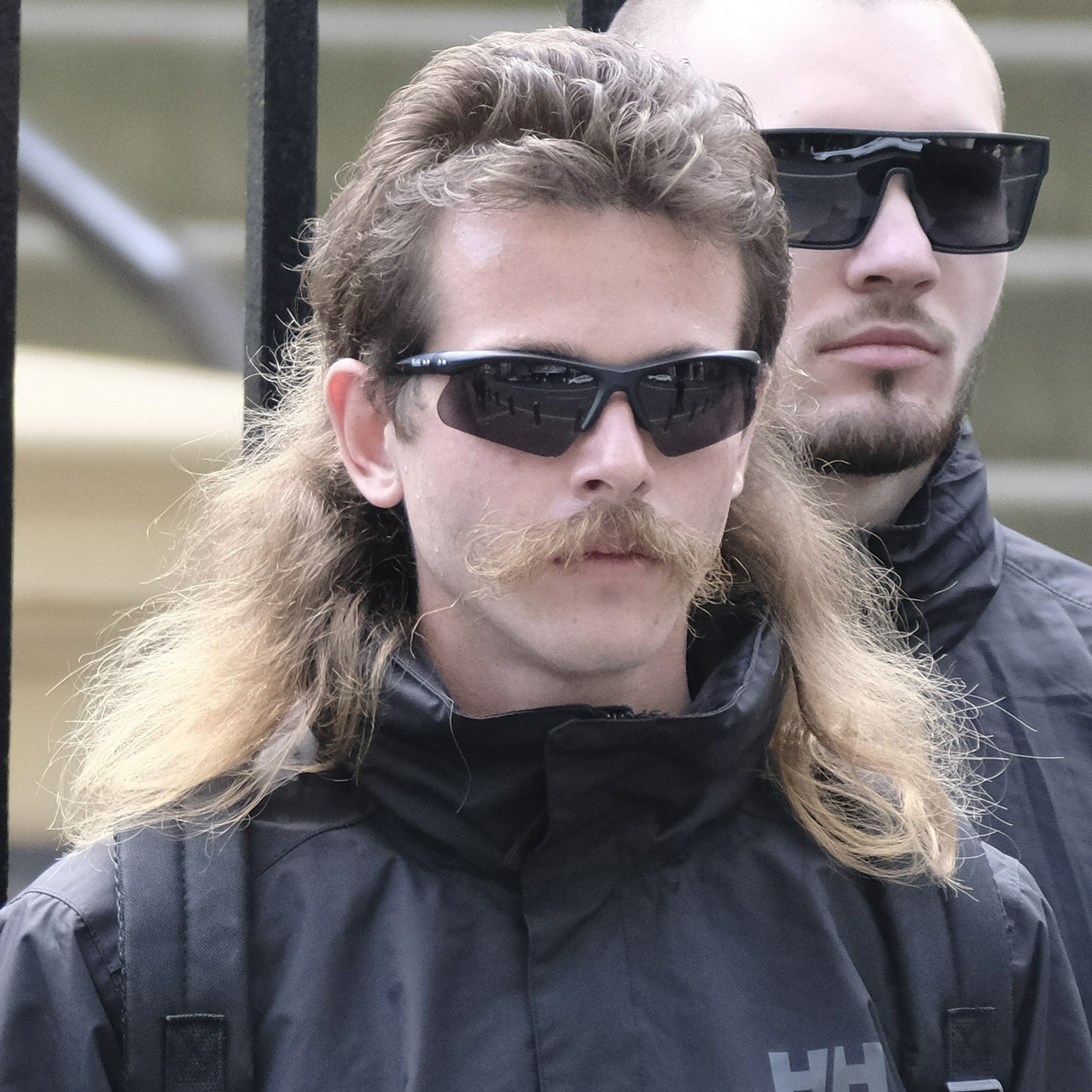 A scrawny man with a frayed brown mullet and very straggly unkempt moustache wears half-frameless black sunglasses