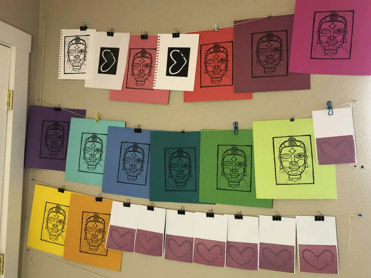 multiple strings of prints hanging against the wall, drying. One set is small cards, with a pink background and purple heart, in phase 2 of a reduction print. The other is a half-awake buddhahead, called "Half-awake Doll", printed on a number of colors of paper. 