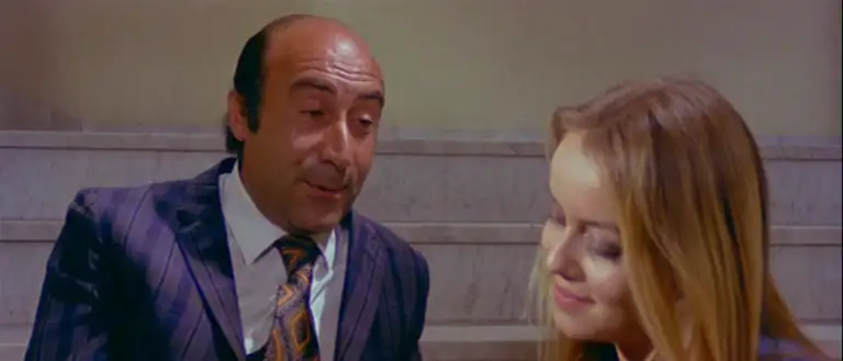 Osvaldo and Loredana sit on steps outside Elivira's apartment and talk. Osvoldo is an older, balding man. He wears a purple suit. Loredana sits closer to the camera, out of focus.
