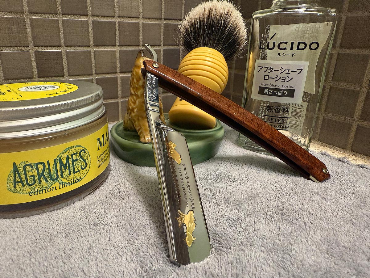 On a gray microfiber towel a straight razor with blade open about 45 degrees leans next to a bottle of aftershave and a jar of shave soap. A shave brush rests bristles-up in a green Wade ceramic pipe rest that features a brown Cairn Terrier.