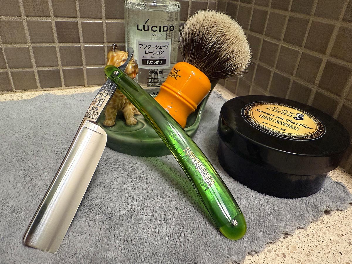 On a gray microfiber towel a straight razor with blade open about 45 degrees leans next to a bottle of aftershave and a jar of shave soap. A shave brush rests bristles-up in a green Wade ceramic pipe rest that features a brown Cairn Terrier.