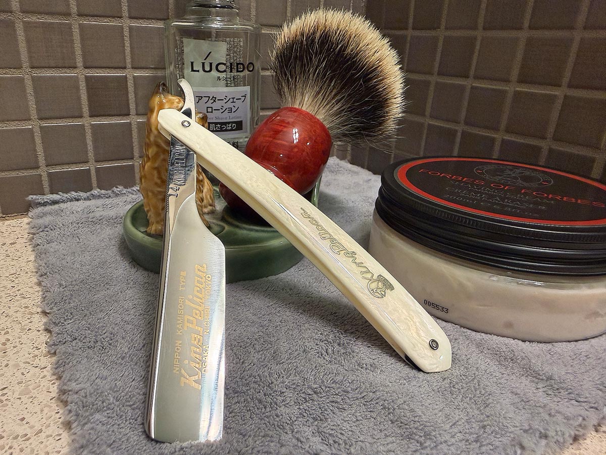 On a gray microfiber towel a straight razor with blade open about 45 degrees leans next to a bottle of aftershave and a jar of shave soap. A shave brush rests bristles-up in a green Wade ceramic pipe rest that features a brown Cairn Terrier.
