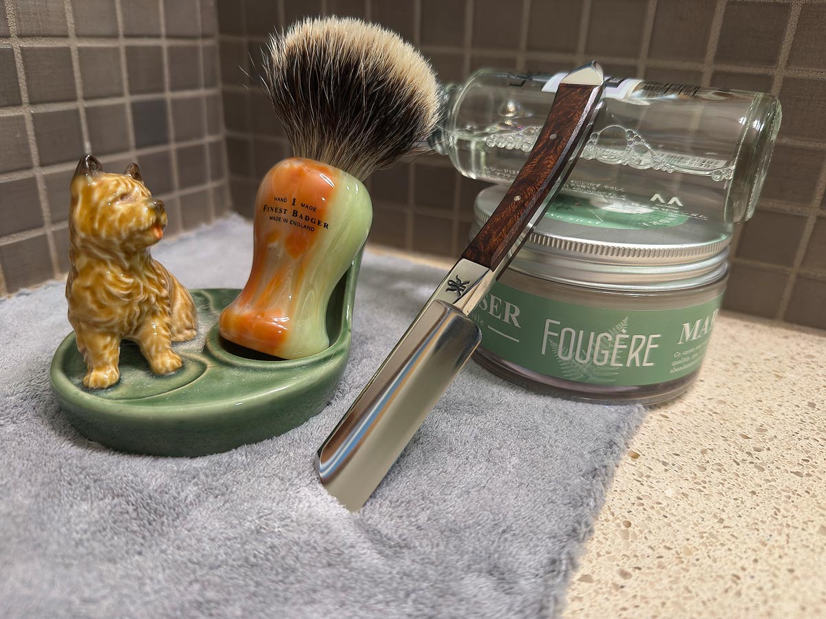 On a gray microfiber towel a straight razor with blade open about 45 degrees leans next to a bottle of aftershave and a jar of shave soap. A shave brush rests bristles-up in a green Wade ceramic pipe rest that features a brown Cairn Terrier.