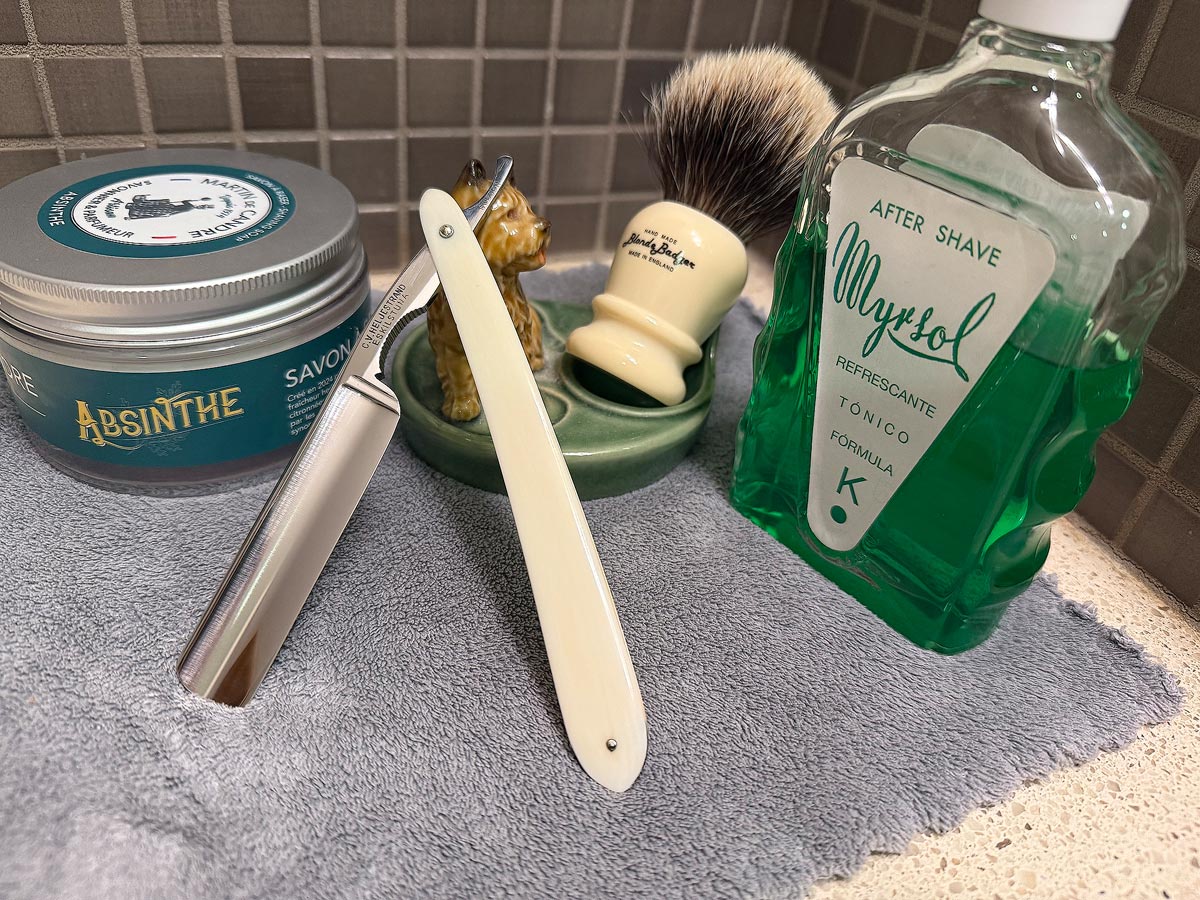 On a gray microfiber towel a straight razor with blade open about 45 degrees leans next to a bottle of aftershave and a jar of shave soap. A shave brush rests bristles-up in a green Wade ceramic pipe rest that features a brown Cairn Terrier.
