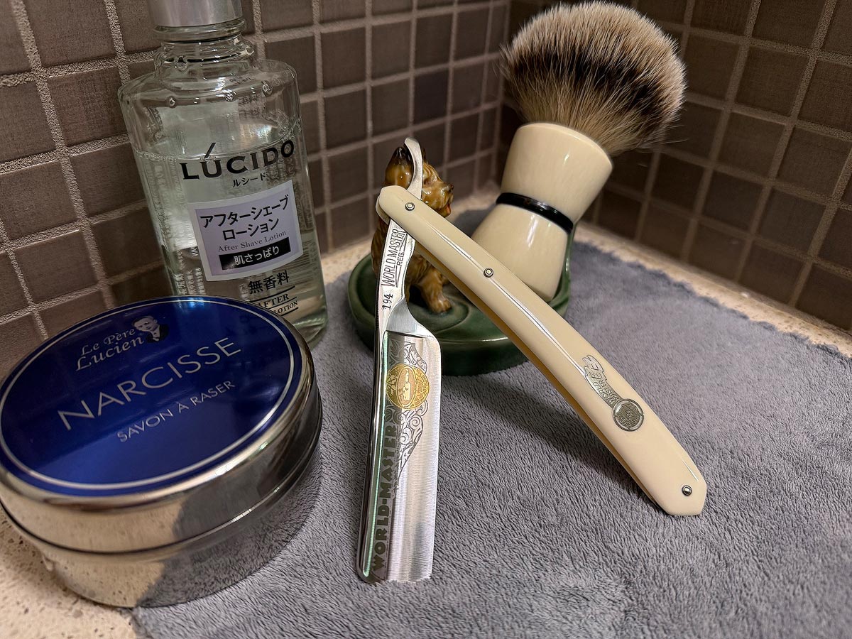 On a gray microfiber towel a straight razor with blade open about 45 degrees leans next to a bottle of aftershave and a jar of shave soap. A shave brush rests bristles-up in a green Wade ceramic pipe rest that features a brown Cairn Terrier.