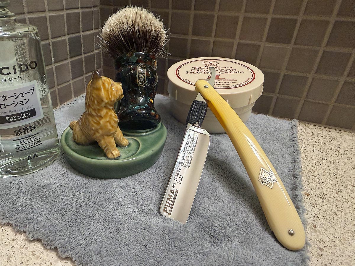 On a gray microfiber towel a straight razor with blade open about 45 degrees leans next to a bottle of aftershave and a jar of shave soap. A shave brush rests bristles-up in a green Wade ceramic pipe rest that features a brown Cairn Terrier.