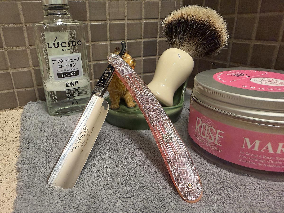 On a gray microfiber towel a straight razor with blade open about 45 degrees leans next to a bottle of aftershave and a jar of shave soap. A shave brush rests bristles-up in a green Wade ceramic pipe rest that features a brown Cairn Terrier.