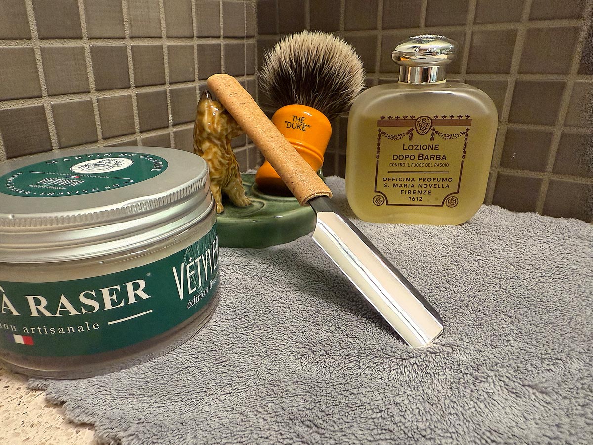 On a gray microfiber towel a straight razor with blade open about 45 degrees leans next to a bottle of aftershave and a jar of shave soap. A shave brush rests bristles-up in a green Wade ceramic pipe rest that features a brown Cairn Terrier.