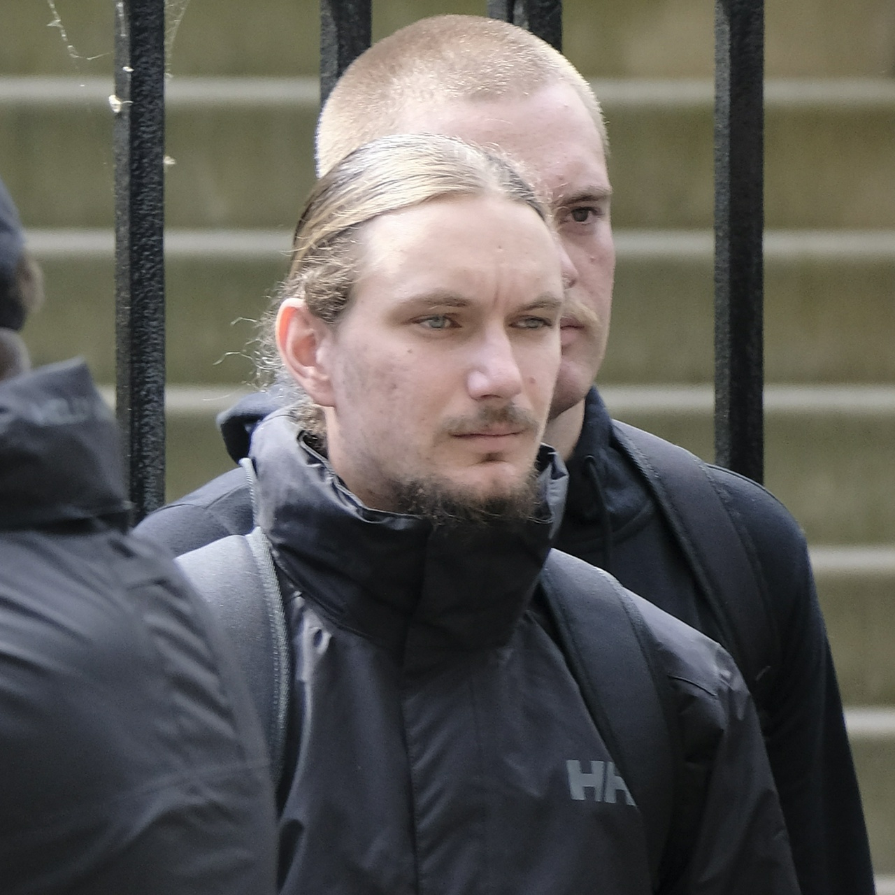 A pockmarked blonde man with long greasy hair, a large tuft on his chin and a mole on his left cheek squints forward