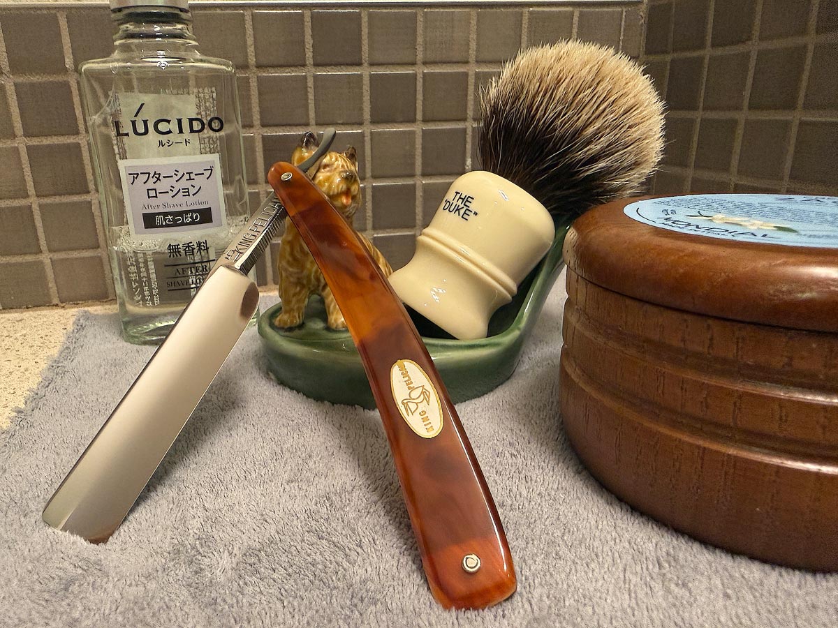 On a gray microfiber towel a straight razor with blade open about 45 degrees leans next to a bottle of aftershave and a jar of shave soap. A shave brush rests bristles-up in a green Wade ceramic pipe rest that features a brown Cairn Terrier.