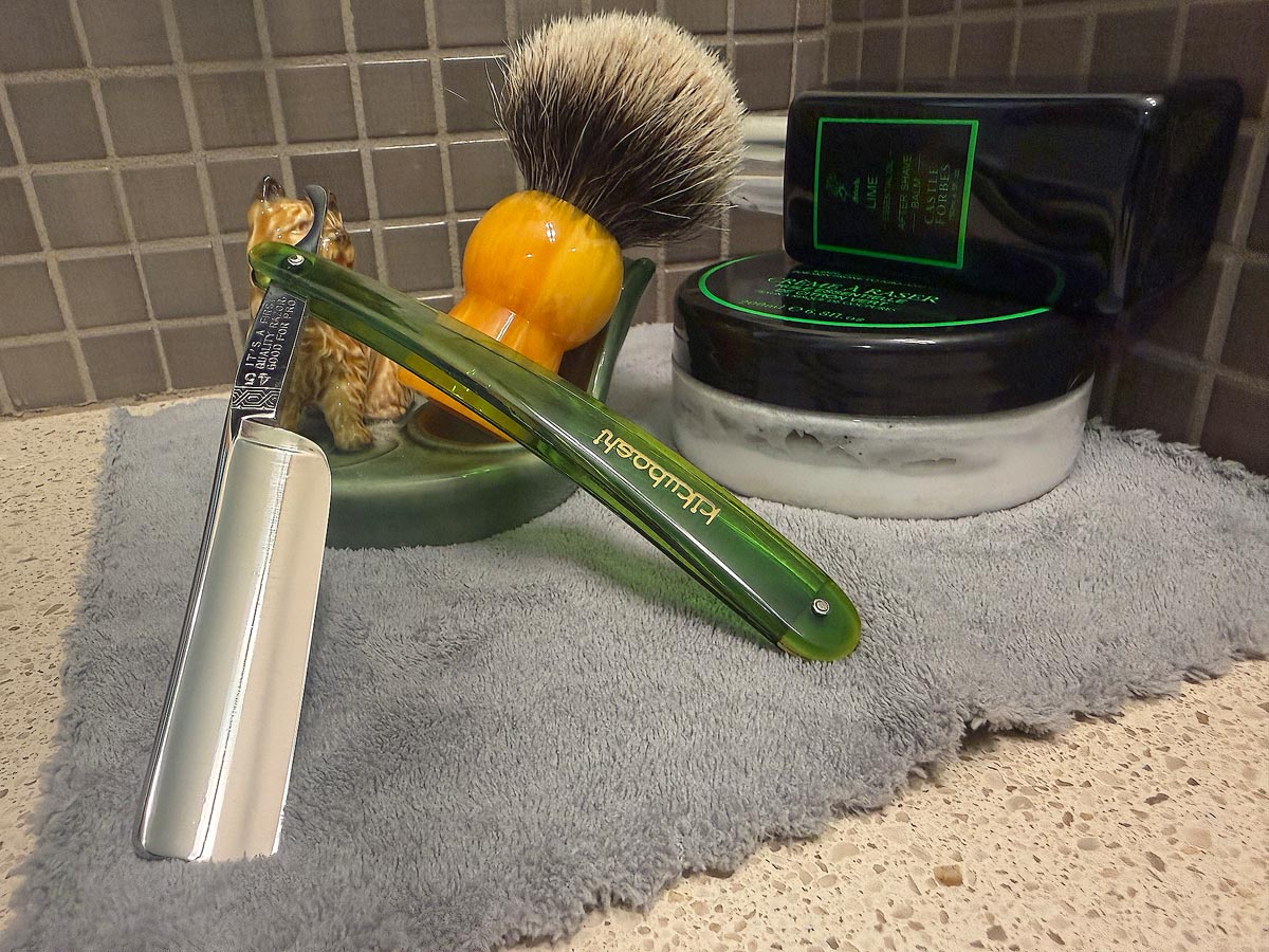 On a gray microfiber towel a straight razor with blade open about 45 degrees leans next to a bottle of aftershave and a jar of shave soap. A shave brush rests bristles-up in a green Wade ceramic pipe rest that features a brown Cairn Terrier.