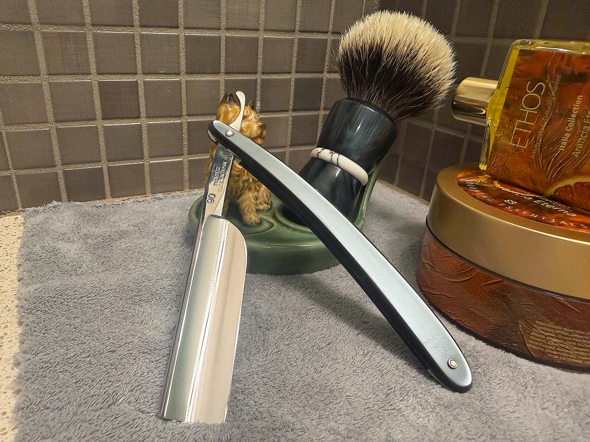 On a gray microfiber towel a straight razor with blade open about 45 degrees leans next to a bottle of aftershave and a jar of shave soap. A shave brush rests bristles-up in a green Wade ceramic pipe rest that features a brown Cairn Terrier.