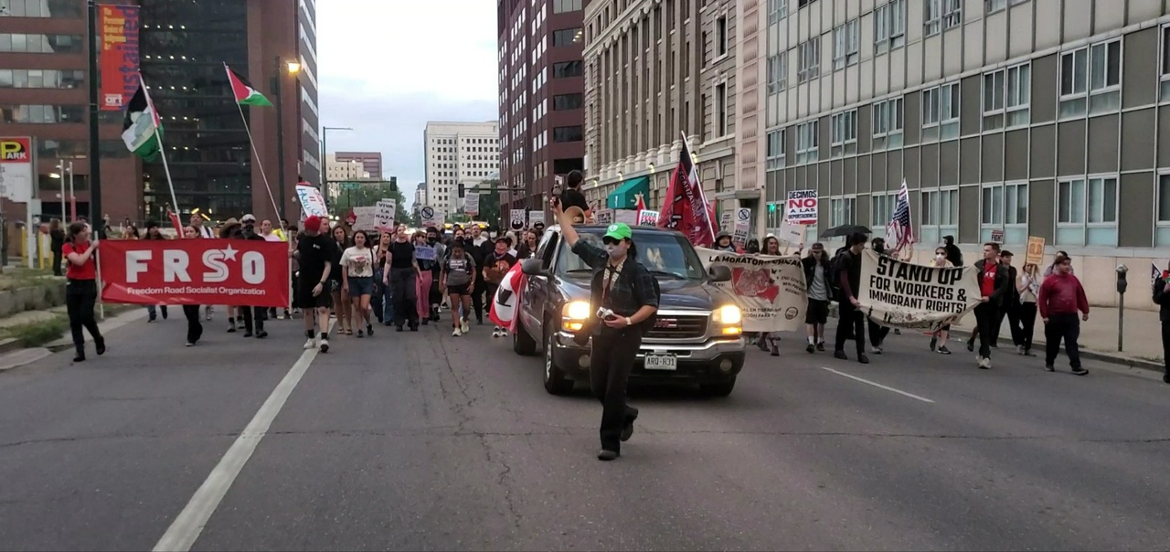 A crowd of over 100 people marches in the streets of Denver carrying Mexican and Palestine flags, led by a vehicle and a protest MC.