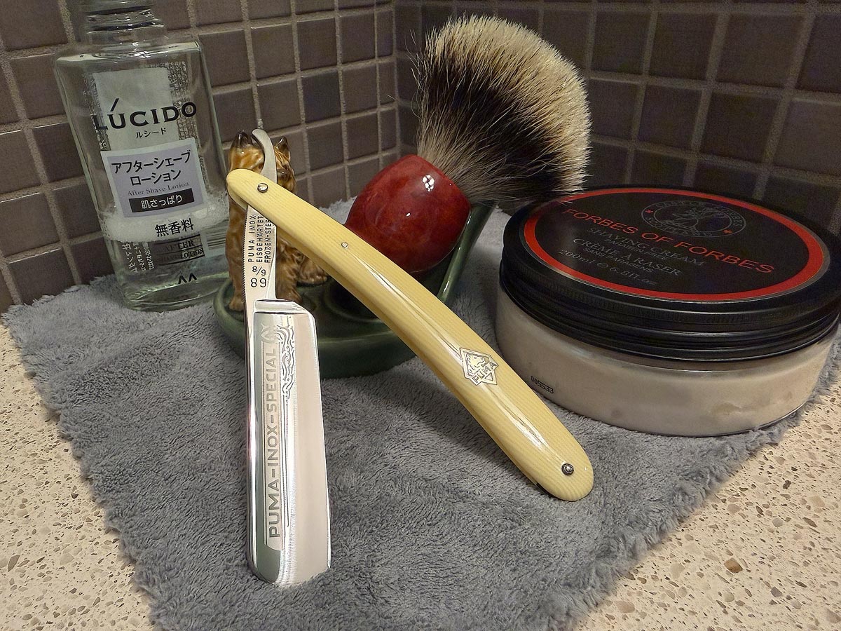 On a gray microfiber towel a straight razor with blade open about 45 degrees leans next to a bottle of aftershave and a jar of shave soap. A shave brush rests bristles-up in a green Wade ceramic pipe rest that features a brown Cairn Terrier.