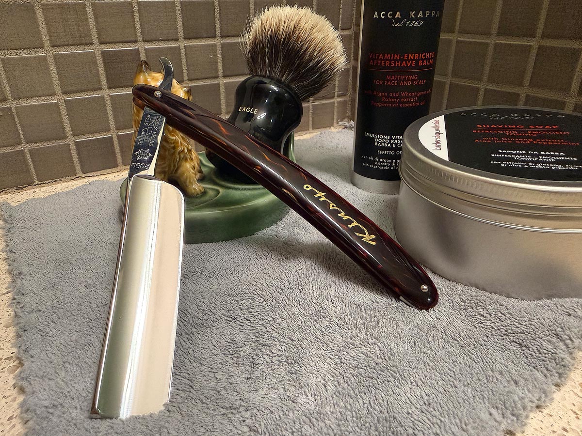 On a gray microfiber towel a straight razor with blade open about 45 degrees leans next to a bottle of aftershave and a jar of shave soap. A shave brush rests bristles-up in a green Wade ceramic pipe rest that features a brown Cairn Terrier.