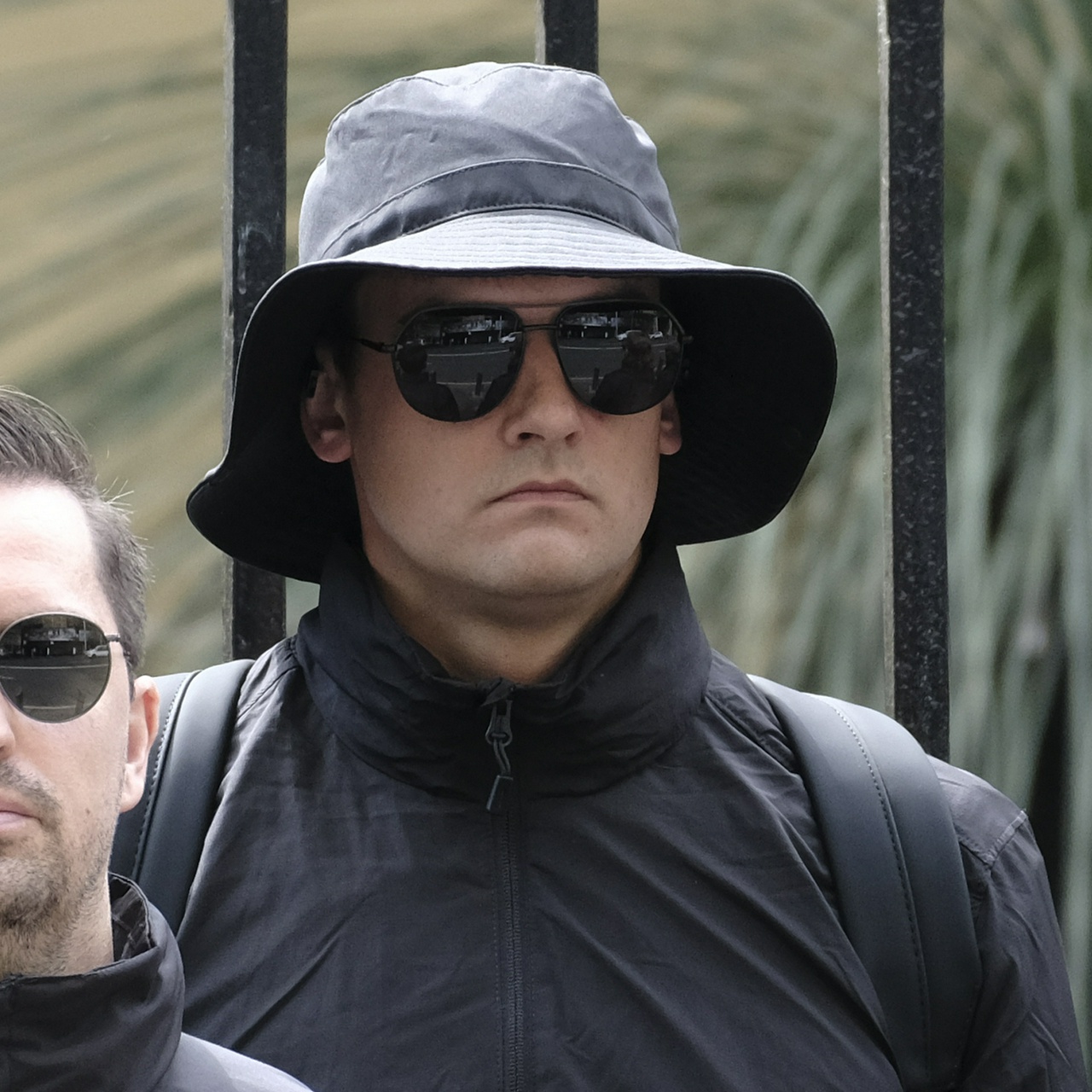 A miserable looking man with a tan and wide neck in a large floppy bucket hat and aviators