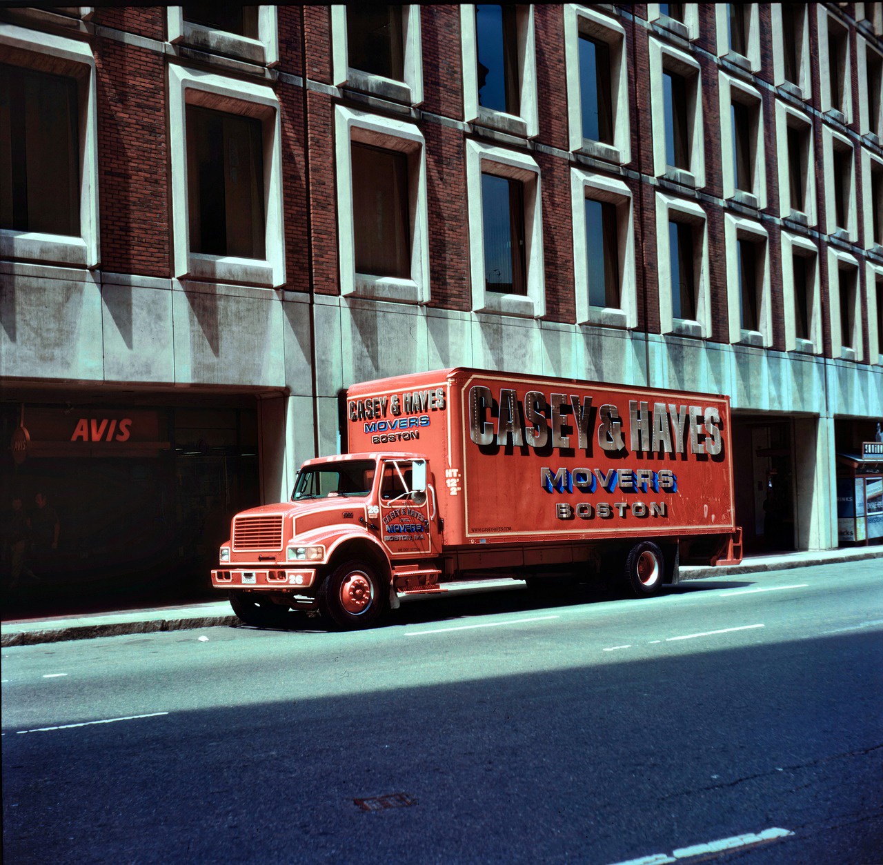 A faded red moving truck in front of a parking garage. The text on the side, door, and front of the truck says Casey & Hayes Movers, Boston.