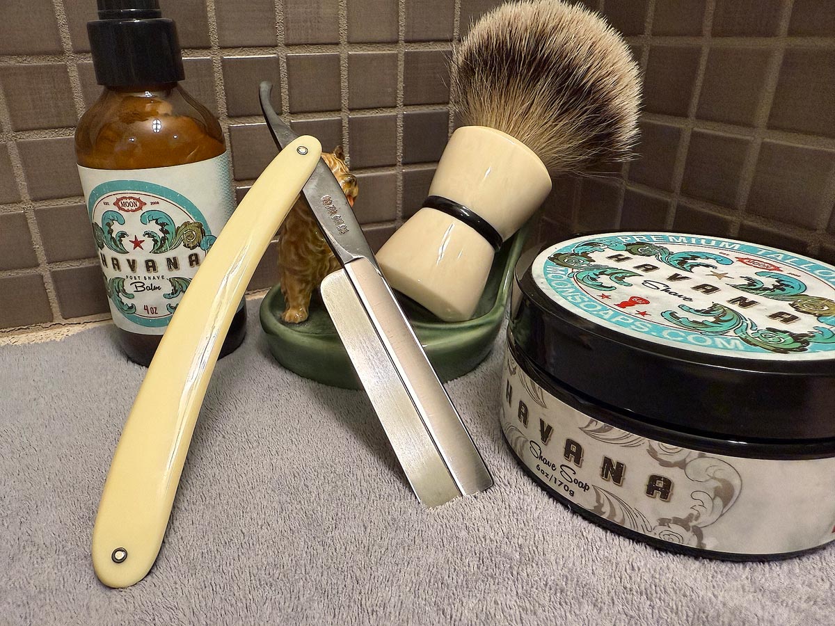 On a gray microfiber towel a straight razor with blade open about 45 degrees leans next to a bottle of aftershave and a jar of shave soap. A shave brush rests bristles-up in a green Wade ceramic pipe rest that features a brown Cairn Terrier.