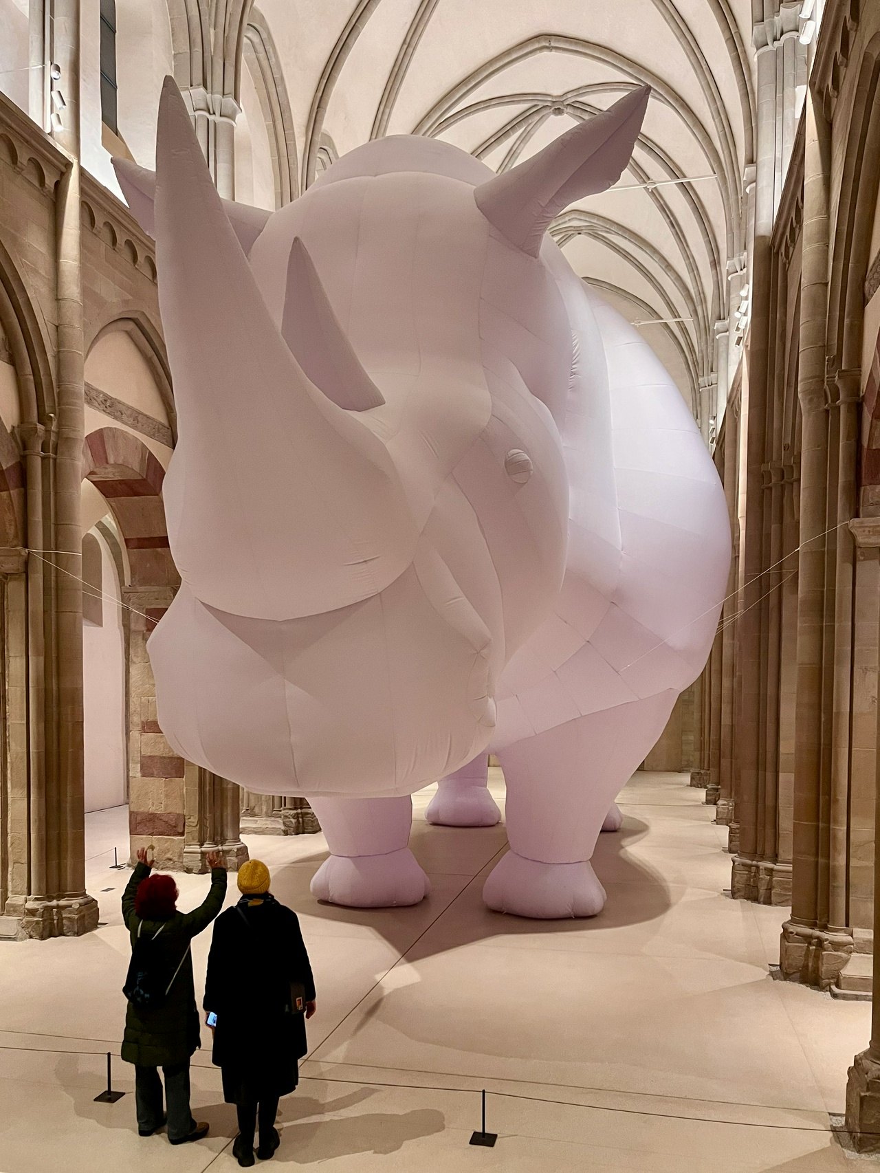 Eine monumentale, weiß aufgeblasene Nashorn-Skulptur füllt nahezu vollständig das Kirchenschiff eines Klosters. Das Tier steht zwischen hohen Steinpfeilern unter einem Gewölbe und wirkt durch seine Größe zugleich weich und überwältigend. Im Vordergrund betrachten zwei Personen mit Mützen die Installation aus der Distanz und heben die Arme, wodurch der Größenunterschied zwischen Mensch und Kunstwerk besonders deutlich wird.