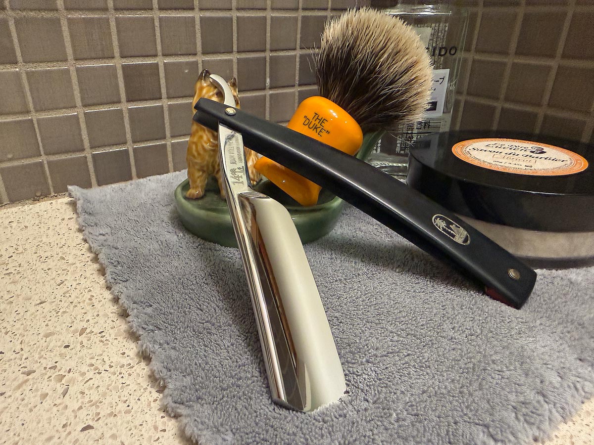 On a gray microfiber towel a straight razor with blade open about 45 degrees leans next to a bottle of aftershave and a jar of shave soap. A shave brush rests bristles-up in a green Wade ceramic pipe rest that features a brown Cairn Terrier.