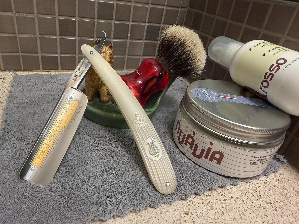 On a gray microfiber towel a straight razor with blade open about 45 degrees leans next to a bottle of aftershave and a jar of shave soap. A shave brush rests bristles-up in a green Wade ceramic pipe rest that features a brown Cairn Terrier.