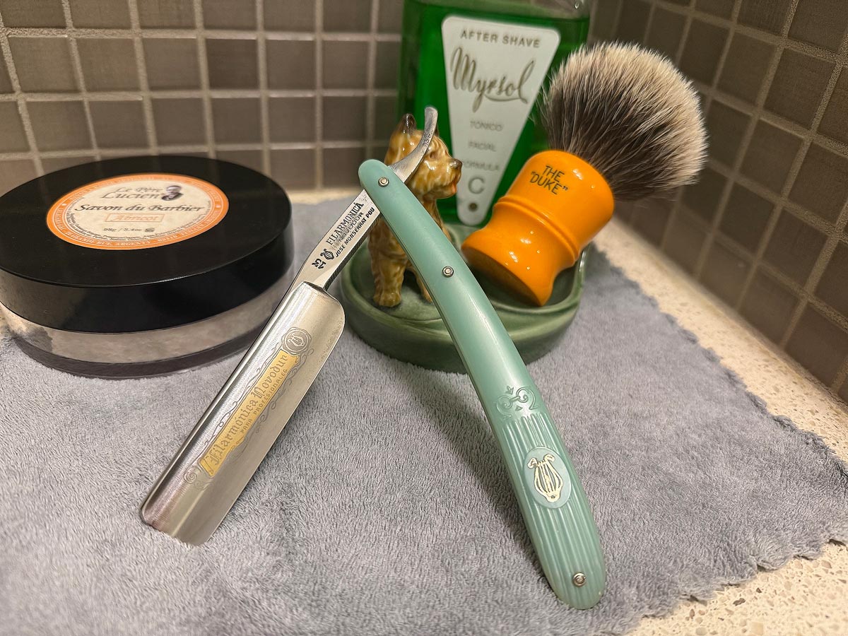On a gray microfiber towel a straight razor with blade open about 45 degrees leans next to a bottle of aftershave and a jar of shave soap. A shave brush rests bristles-up in a green Wade ceramic pipe rest that features a brown Cairn Terrier.