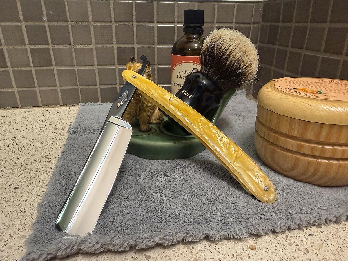 On a gray microfiber towel a straight razor with blade open about 45 degrees leans next to a bottle of aftershave and a jar of shave soap. A shave brush rests bristles-up in a green Wade ceramic pipe rest that features a brown Cairn Terrier.
