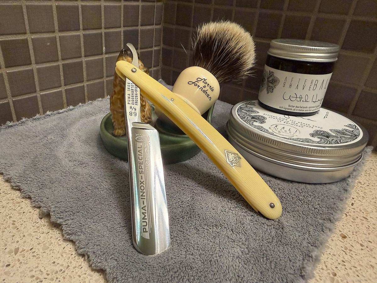 On a gray microfiber towel a straight razor with blade open about 45 degrees leans next to a bottle of aftershave and a jar of shave soap. A shave brush rests bristles-up in a green Wade ceramic pipe rest that features a brown Cairn Terrier.
