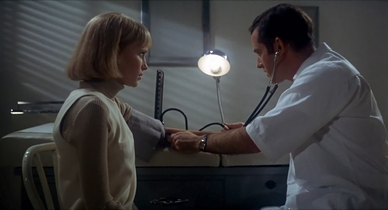 Movie still from Rosemary's Baby, with Mia Farrow and Charles Grodin seen in profile, facing each other as the doctor takes her blood pressure
