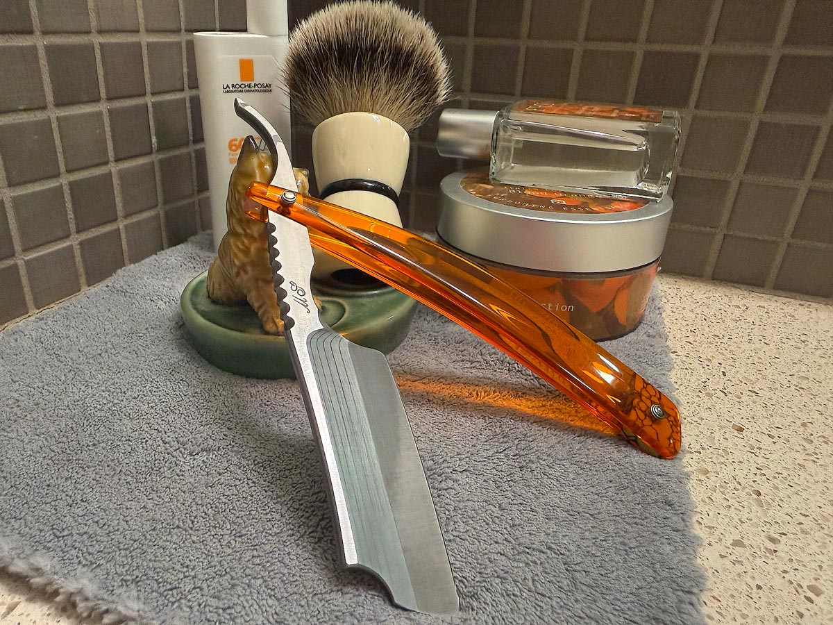 On a gray microfiber towel a straight razor with blade open about 45 degrees leans next to a bottle of aftershave and a jar of shave soap. A shave brush rests bristles-up in a green Wade ceramic pipe rest that features a brown Cairn Terrier.