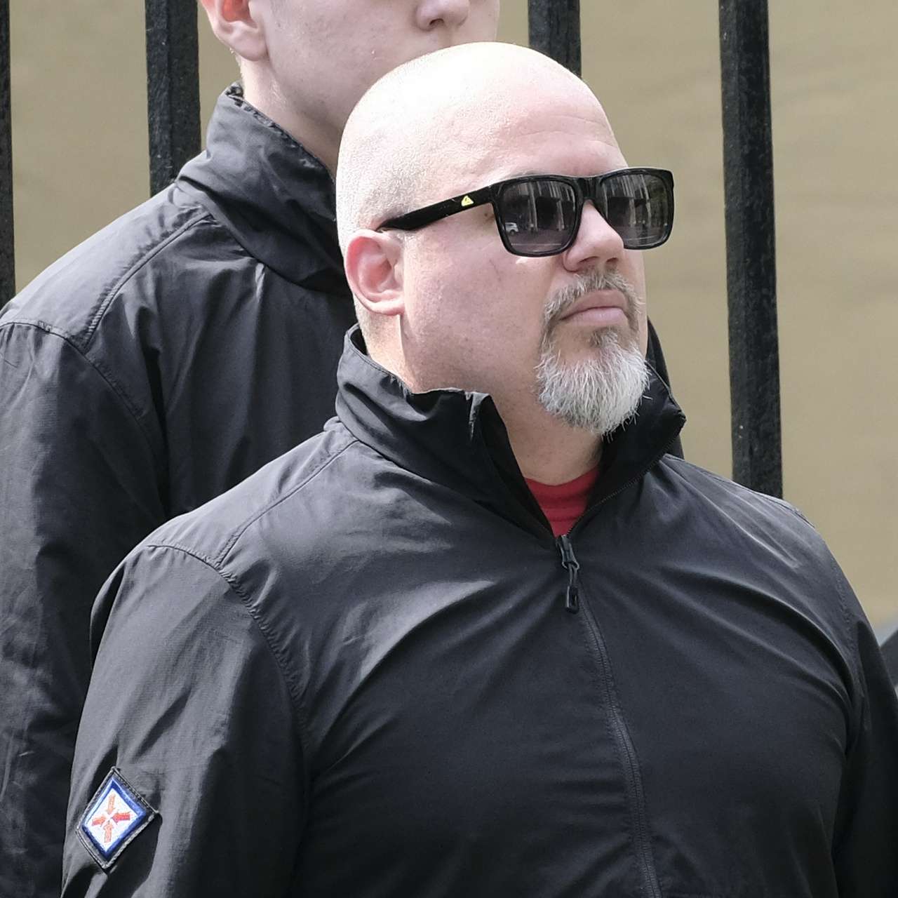 An old bald man with a grey goatee and black sunglasses, his jacket too small to zip up over his bright red shirt underneath