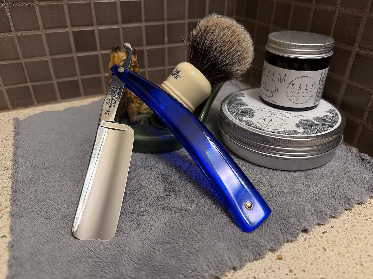 On a gray microfiber towel a straight razor with blade open about 45 degrees leans next to a bottle of aftershave and a jar of shave soap. A shave brush rests bristles-up in a green Wade ceramic pipe rest that features a brown Cairn Terrier.