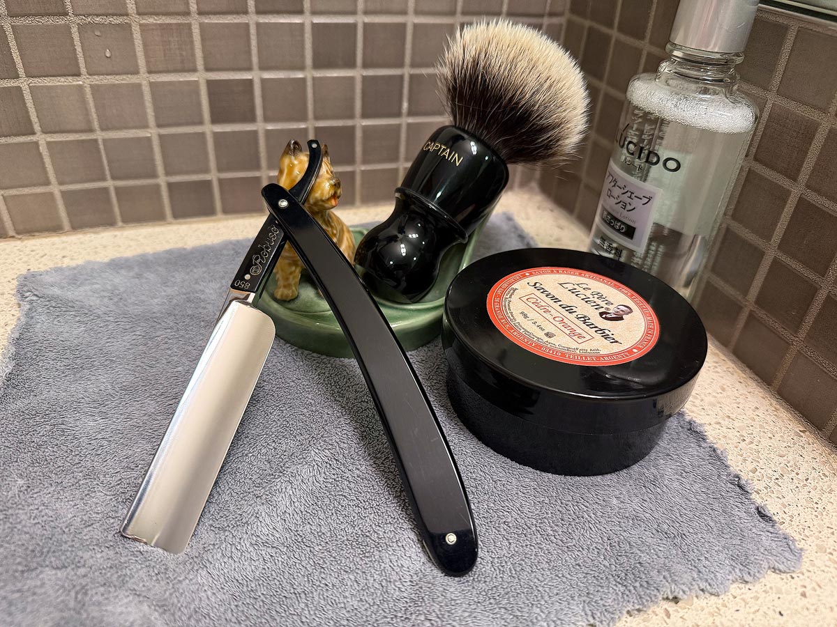 On a gray microfiber towel a straight razor with blade open about 45 degrees leans next to a bottle of aftershave and a jar of shave soap. A shave brush rests bristles-up in a green Wade ceramic pipe rest that features a brown Cairn Terrier.