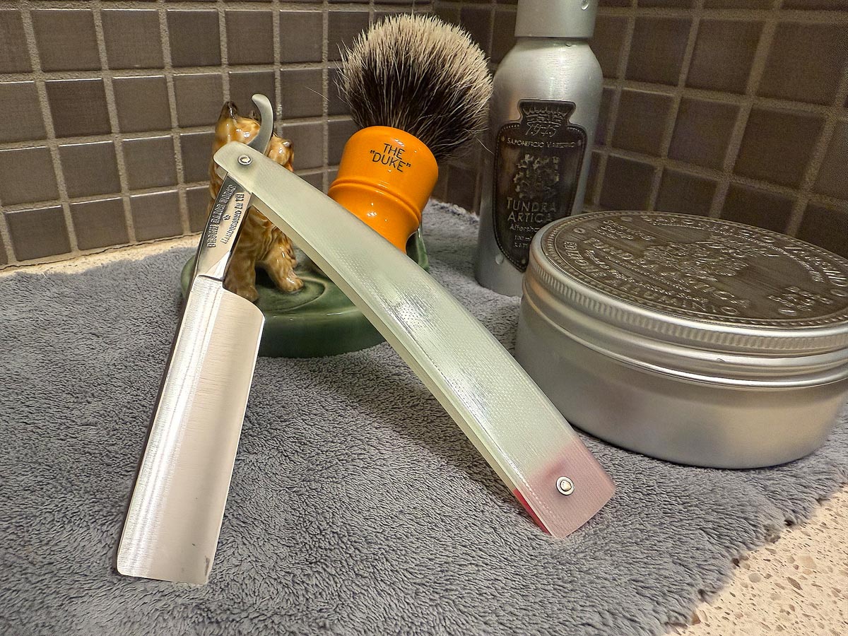On a gray microfiber towel a straight razor with blade open about 45 degrees leans next to a bottle of aftershave and a jar of shave soap. A shave brush rests bristles-up in a green Wade ceramic pipe rest that features a brown Cairn Terrier.