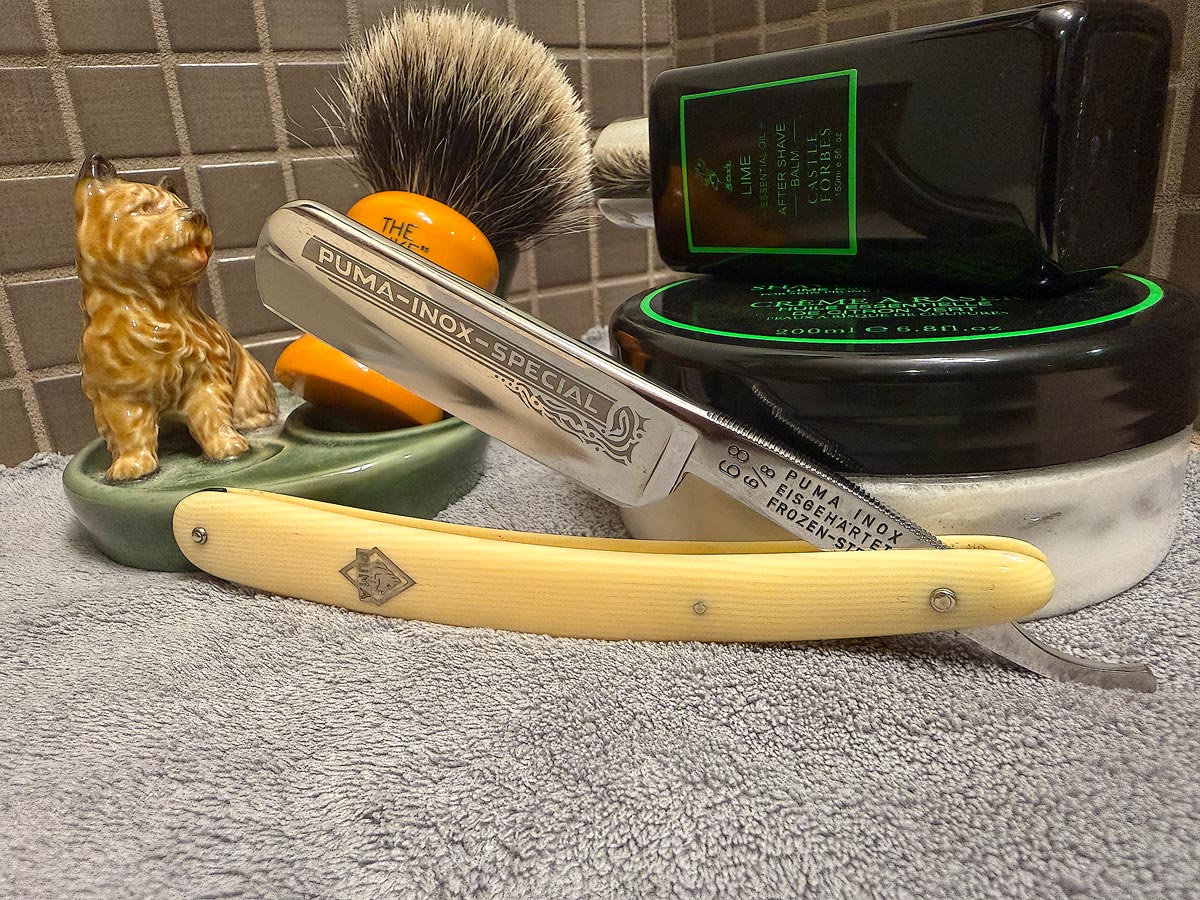 On a gray microfiber towel a straight razor with blade open about 45 degrees leans next to a bottle of aftershave and a jar of shave soap. A shave brush rests bristles-up in a green Wade ceramic pipe rest that features a brown Cairn Terrier.